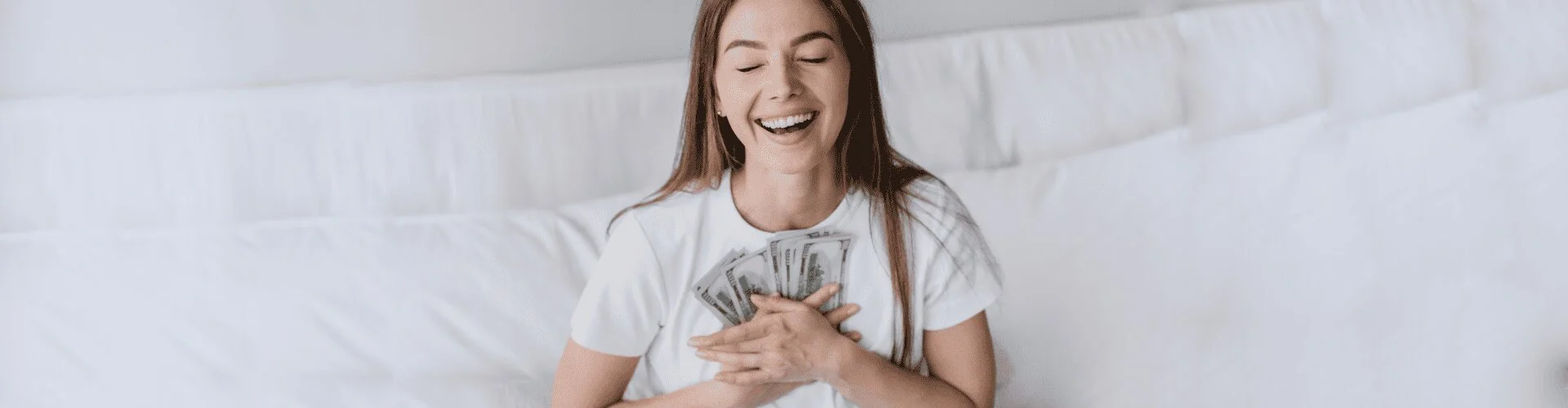 Happy woman holding money and smiling on a bed.