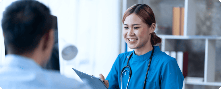 Young female nurse in blue scrubs with a stethoscope, smiling and holding a clipboard while talking to a patient.