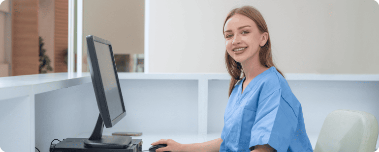 Female nurse in blue scrubs smiling at a computer in a hospital reception area.
