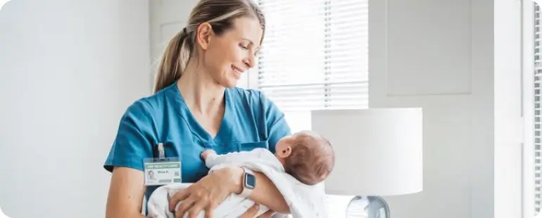 Smiling nurse in blue scrubs holding a newborn in hospital room